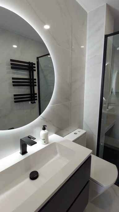Baño moderno con lavabo blanco, espejo circular y detalles en gris y negro.