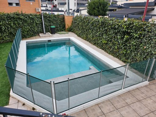 Piscina con cercado de vidrio en un jardín, rodeada de seto verde.