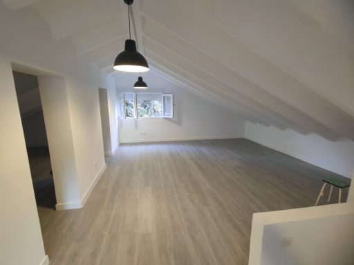 Espacio interior amplio con techo a dos aguas, paredes blancas y suelos de madera.